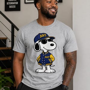 Op de afbeelding: Een grijs t-shirt met een cartoon Snoopy-personage met een zonnebril, een blauw en gouden baseballpet en een bijpassende jas met de letters "A&T". De afbeelding is van een lachende persoon.