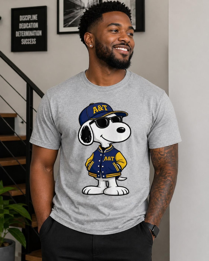 Op de afbeelding: Een grijs t-shirt met een cartoon Snoopy-personage met een zonnebril, een blauw en gouden baseballpet en een bijpassende jas met de letters "A&T". De afbeelding is van een lachende persoon.