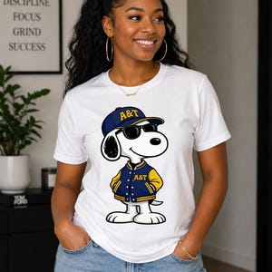 Op de afbeelding: Wit T-shirt met een cartoonhond met een zonnebril, een blauwe pet met de tekst "A&T" en een blauwgele jas. De hond staat in een ontspannen pose. Het shirt wordt gecombineerd met een blauwe jeans.