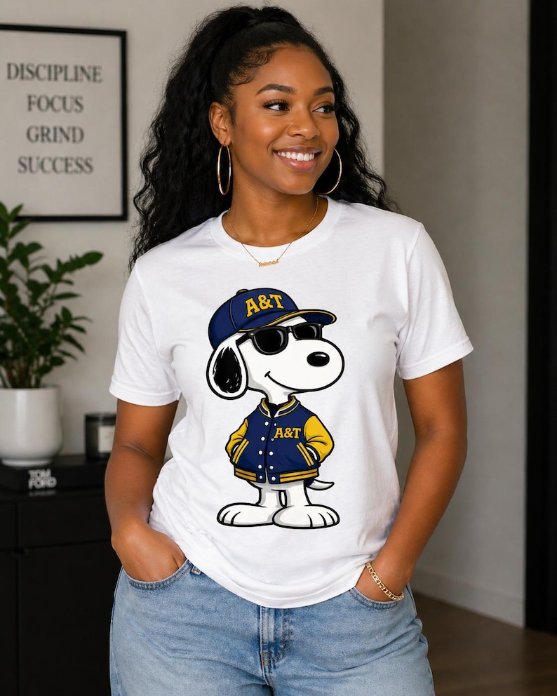 Op de afbeelding: Wit T-shirt met een cartoonhond met een zonnebril, een blauwe pet met de tekst "A&T" en een blauwgele jas. De hond staat in een ontspannen pose. Het shirt wordt gecombineerd met een blauwe jeans.
