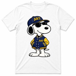 Op de afbeelding: Wit T-shirt met een cartoonhond die een marineblauwe baseballcap en jas met gele accenten draagt. Op de pet en jas staan de letters "A&T". De hond draagt ook een zwarte zonnebril.