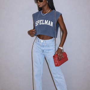 May include: A navy blue sleeveless cropped t-shirt with the word SPELMAN printed in white block letters across the chest, paired with light wash blue denim jeans featuring vertical seam detailing and a red quilted shoulder bag with a chain strap.