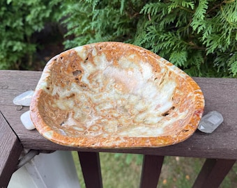 May include: A natural stone bowl with a unique pattern of tan, brown, and cream colors. The bowl has a smooth, polished interior and a rougher exterior. Two clear quartz crystals are placed near the bowl. The bowl is on a wooden surface.