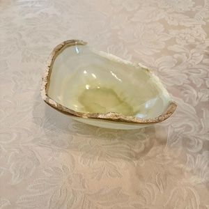 May include: A translucent, light green onyx bowl with a rough, natural edge. The bowl has a smooth interior and a slightly irregular shape, showcasing the natural stone's unique patterns and colors. The rim has a textured, light brown edge.