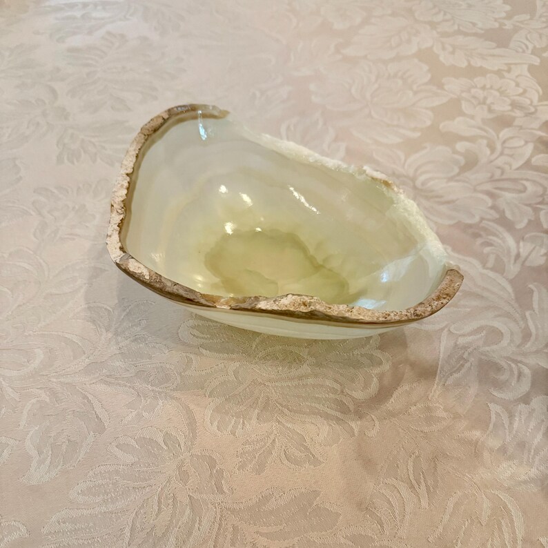 May include: A translucent, light green onyx bowl with a rough, natural edge. The bowl has a smooth interior and a slightly irregular shape, showcasing the natural stone's unique patterns and colors. The rim has a textured, light brown edge.