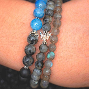 May include: Three layered bracelets with black, gray, and blue beads. The top layer has two light blue beads. The middle layer has a silver charm.