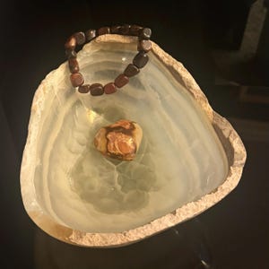 May include: A decorative, light green onyx bowl with a rough, beige rim. A dark brown beaded bracelet rests in the bowl, along with a small, heart-shaped stone with brown and tan patterns. The bowl sits on a dark surface.