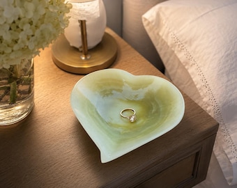 May include: A heart-shaped dish with a smooth, translucent finish in shades of pale green and cream. The dish features a swirling, marbled pattern throughout its surface and contains a single gold-toned ring with a clear stone in its center.