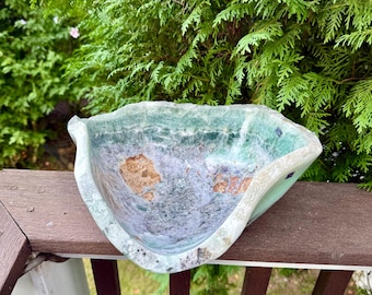 May include: A handcrafted, natural stone bowl with a unique, organic shape. The bowl features a blend of green, blue, and brown hues, with a textured surface. The bowl is displayed outdoors, showcasing its natural beauty and craftsmanship.