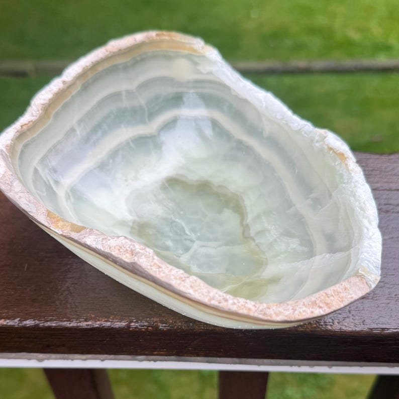 May include: A light green and white onyx bowl with a natural, uneven edge. The bowl has a layered, translucent appearance, with bands of color. The rim has a rough, textured, light brown edge. The bowl is on a dark brown surface.