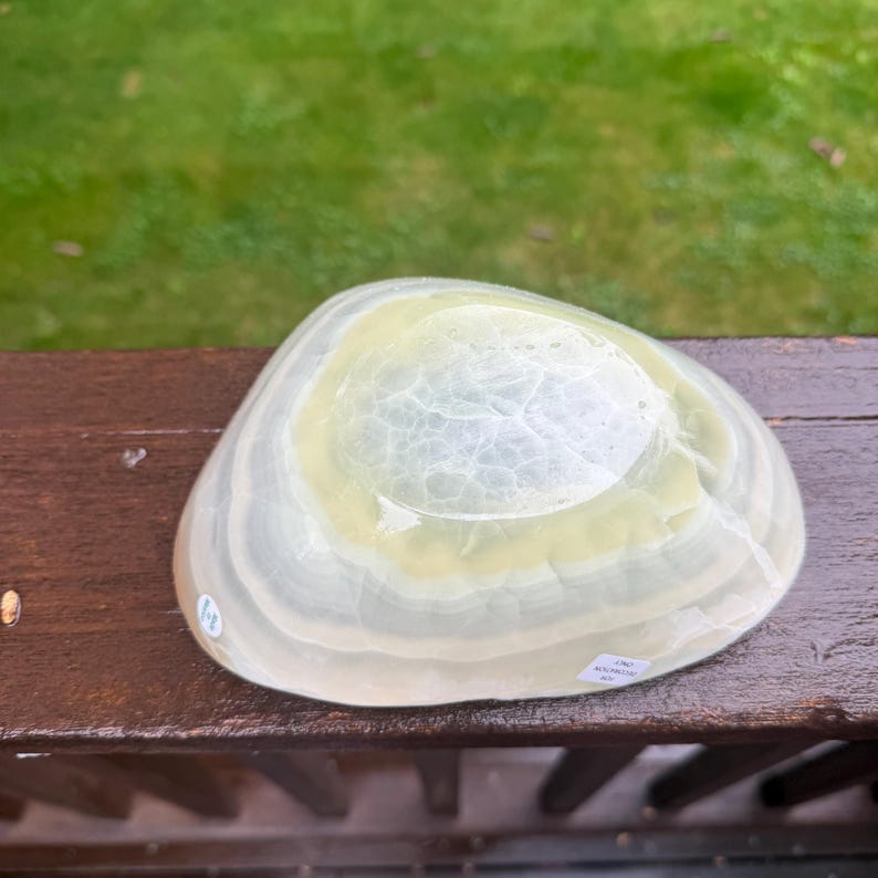 May include: A translucent, oval-shaped decorative bowl with a light green and white layered design. The bowl has a smooth, polished surface and a shallow, rounded interior. The bowl is sitting on a wooden surface.
