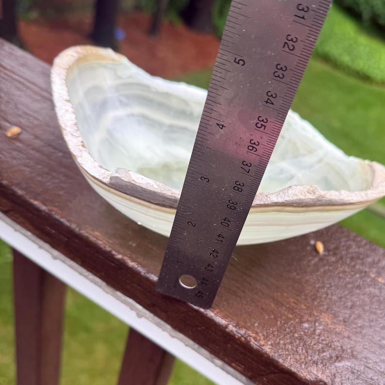 May include: A light green and beige onyx bowl with a rough, natural edge. The bowl is measured with a ruler, showing a width of approximately 5 inches. The bowl's interior has a layered, translucent appearance.