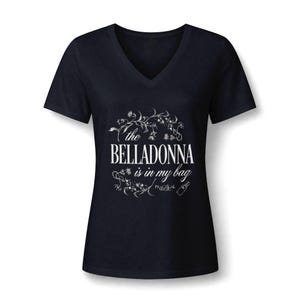 May include: A black V-neck t-shirt featuring white graphic text that reads "the BELLADONNA is in my bag" surrounded by botanical illustrations of flowers and vines, with the small text "PRACTICAL MAGIC" and a bottle icon at the bottom.