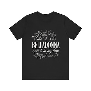 Peut inclure: Un t-shirt noir à manches courtes avec le texte blanc the BELLADONNA is in my bag et PRACTICAL MAGIC. Le motif comprend une illustration botanique blanche de vignes et de fleurs entourant le texte ainsi qu'une petite icône de bouteille.