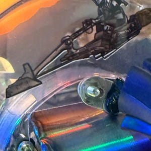 Puede incluir: Primer plano de un detalle de m&aacute;quina de pinball con una ilustraci&oacute;n en blanco y negro de un personaje montado en una speeder bike. La imagen tiene una superficie reflectante e iridiscente con tonos naranja, azul y verde.