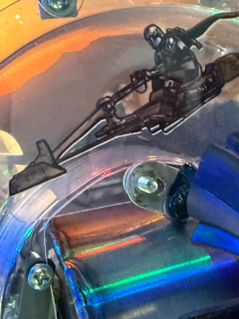 Puede incluir: Primer plano de un detalle de m&aacute;quina de pinball con una ilustraci&oacute;n en blanco y negro de un personaje montado en una speeder bike. La imagen tiene una superficie reflectante e iridiscente con tonos naranja, azul y verde.