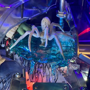 May include: A white, multi-legged spider figurine positioned on a translucent, textured blue ice-like platform. The spider has a segmented body and long, thin legs. Below the blue platform, a black and white illustration of icicles is visible.