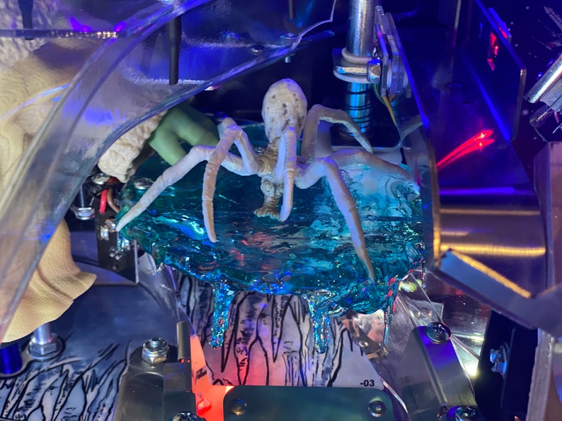 May include: A white, multi-legged spider figurine positioned on a translucent, textured blue ice-like platform. The spider has a segmented body and long, thin legs. Below the blue platform, a black and white illustration of icicles is visible.