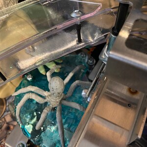 May include: A light gray spider figurine with eight legs positioned on a translucent blue textured base. The spider is situated beneath a clear plastic ramp with metal hardware, including screws and a bracket, visible in the pinball machine.