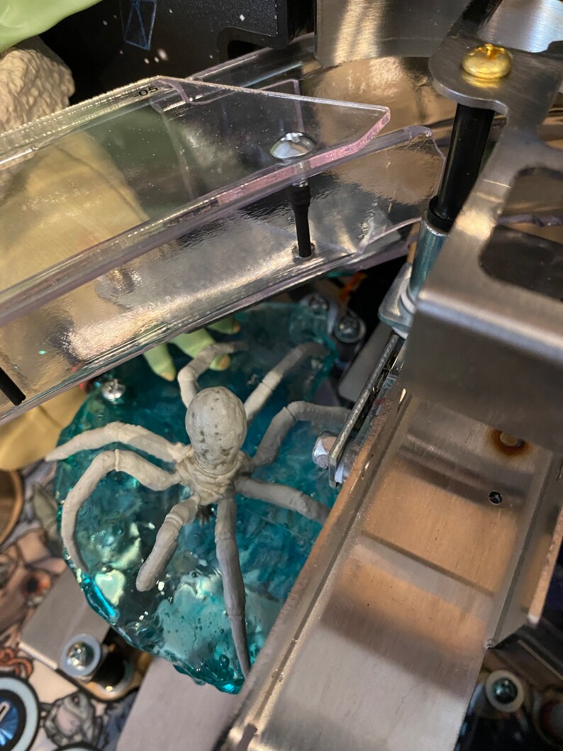 May include: A light gray spider figurine with eight legs positioned on a translucent blue textured base. The spider is situated beneath a clear plastic ramp with metal hardware, including screws and a bracket, visible in the pinball machine.