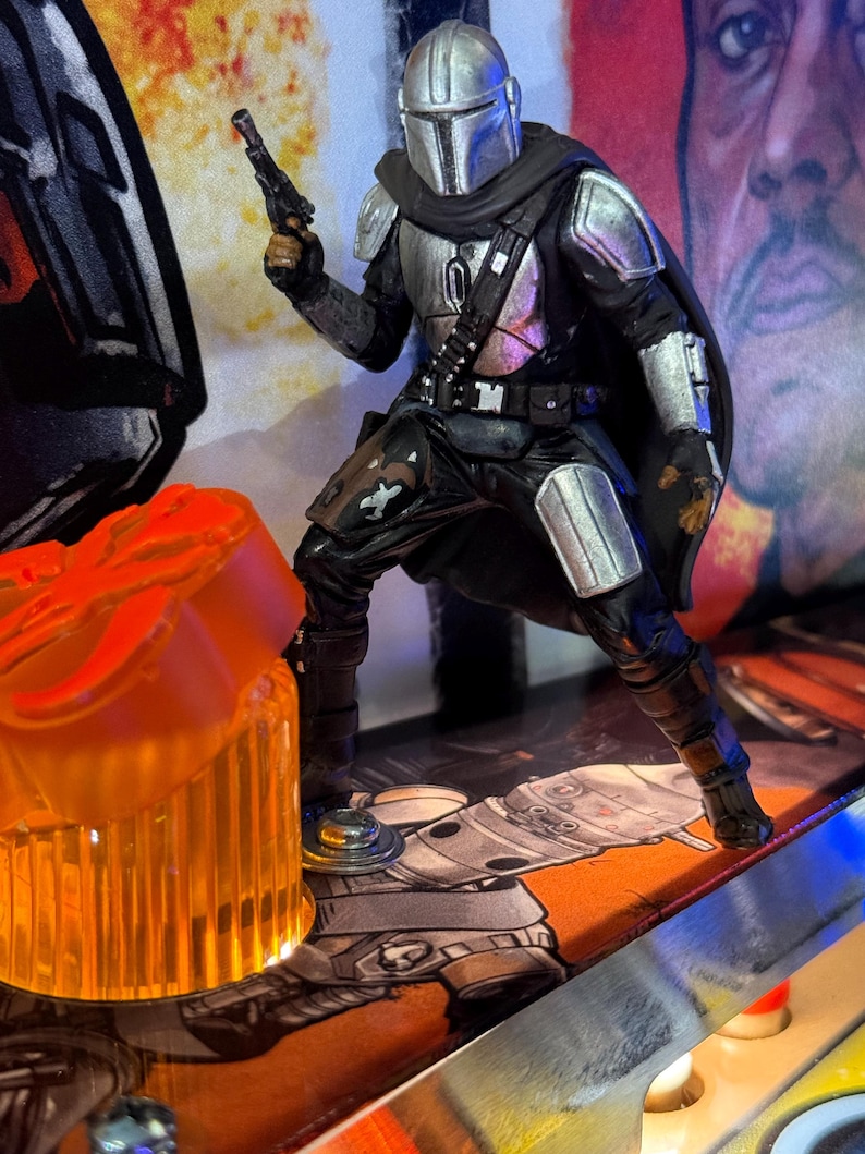 Puede incluir: Figura de acci&oacute;n detallada de The Mandalorian, vestido con armadura plateada y una capa negra, sosteniendo una pistola bl&aacute;ster. La figura est&aacute; posada en una m&aacute;quina de pinball, con una l&aacute;mpara naranja brillante en primer plano y un fondo tem&aacute;tico de Star Wars.