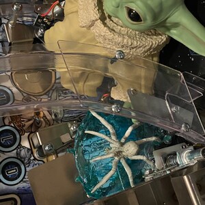 May include: A green creature figurine with large dark eyes and a tan textured robe positioned above a translucent blue ice-like base featuring a white spider-like creature. The scene is set within a pinball machine playfield with metal components.