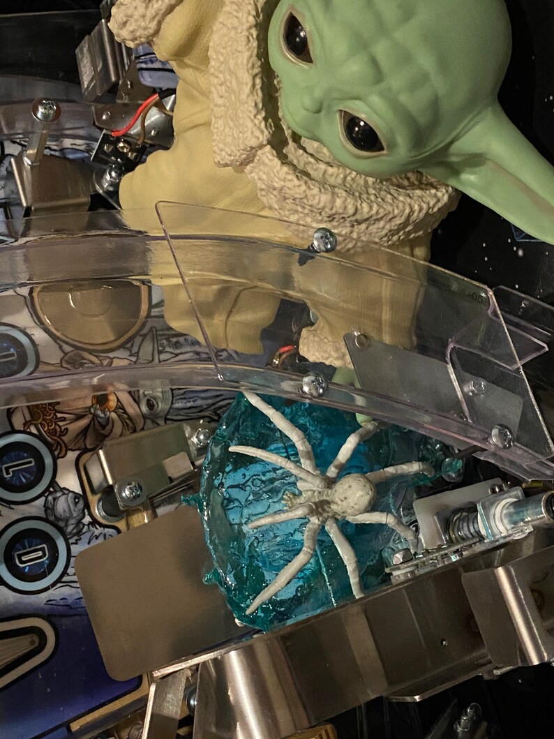 May include: A green creature figurine with large dark eyes and a tan textured robe positioned above a translucent blue ice-like base featuring a white spider-like creature. The scene is set within a pinball machine playfield with metal components.