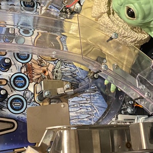 May include: A pinball machine playfield featuring a green creature figurine with large black eyes wearing a tan coat with a fuzzy collar. The playfield surface displays blue and white graphics with the text "COLD" and an illustration of the creature.