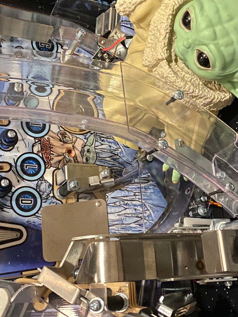 May include: A pinball machine playfield featuring a green creature figurine with large black eyes wearing a tan coat with a fuzzy collar. The playfield surface displays blue and white graphics with the text "COLD" and an illustration of the creature.