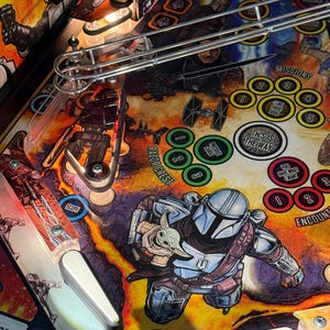 Puede incluir: Una vista detallada del campo de juego de una m&aacute;quina de pinball con personajes de una popular serie de ciencia ficci&oacute;n. La obra de arte incluye im&aacute;genes de un cazarrecompensas y un peque&ntilde;o alien&iacute;gena verde. El campo de juego tiene varios objetivos, rampas y el texto "THIS IS THE WAY".