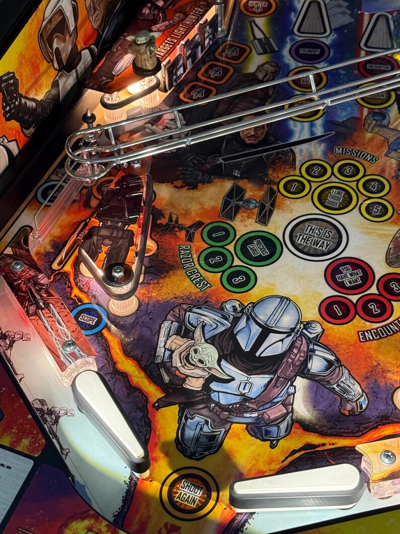 Puede incluir: Una vista detallada del campo de juego de una m&aacute;quina de pinball con personajes de una popular serie de ciencia ficci&oacute;n. La obra de arte incluye im&aacute;genes de un cazarrecompensas y un peque&ntilde;o alien&iacute;gena verde. El campo de juego tiene varios objetivos, rampas y el texto "THIS IS THE WAY".
