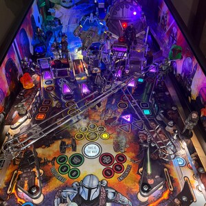 Puede incluir: Una m&aacute;quina de pinball con tem&aacute;tica de Star Wars que presenta personajes como The Mandalorian y Grogu. El campo de juego tiene varias rampas, parachoques y objetivos, con el texto "THIS IS THE WAY" visible. La m&aacute;quina est&aacute; brillantemente iluminada con luces moradas y azules.