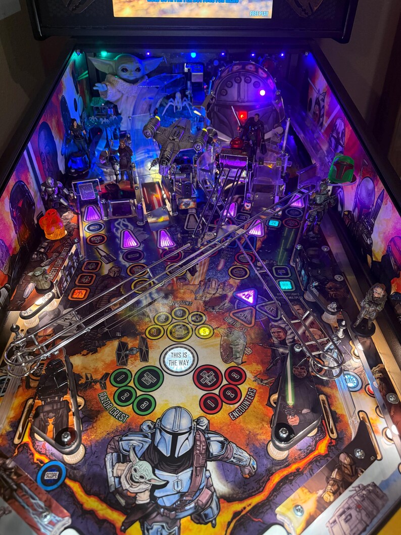 Puede incluir: Una m&aacute;quina de pinball con tem&aacute;tica de Star Wars que presenta personajes como The Mandalorian y Grogu. El campo de juego tiene varias rampas, parachoques y objetivos, con el texto "THIS IS THE WAY" visible. La m&aacute;quina est&aacute; brillantemente iluminada con luces moradas y azules.