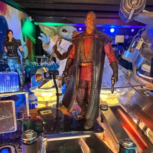 Puede incluir: Una m&aacute;quina de pinball con tem&aacute;tica de Star Wars que presenta varias figuras de personajes. La figura central es un hombre con un atuendo rojo y negro, sosteniendo un arma. Otras figuras incluyen una mujer con armadura negra y una figura de Grogu. La m&aacute;quina tiene un dise&ntilde;o futurista.