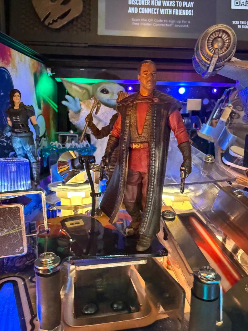 Puede incluir: Una m&aacute;quina de pinball con tem&aacute;tica de Star Wars que presenta varias figuras de personajes. La figura central es un hombre con un atuendo rojo y negro, sosteniendo un arma. Otras figuras incluyen una mujer con armadura negra y una figura de Grogu. La m&aacute;quina tiene un dise&ntilde;o futurista.