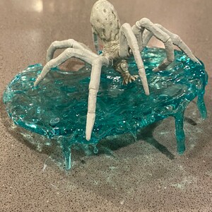May include: A light gray spider figurine with a textured, bulbous body and eight articulated legs, positioned on a translucent, textured teal base that mimics the appearance of rippling water with dripping edges extending downward.