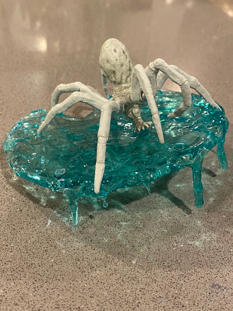 May include: A light gray spider figurine with a textured, bulbous body and eight articulated legs, positioned on a translucent, textured teal base that mimics the appearance of rippling water with dripping edges extending downward.