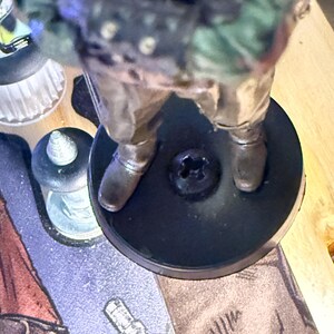 Puede incluir: Una figura en miniatura de un personaje con un atuendo de estilo militar, de pie sobre una base circular negra. La figura tiene una pintura detallada con tonos de verde, marr&oacute;n y plata. La base tiene un tornillo en el centro.