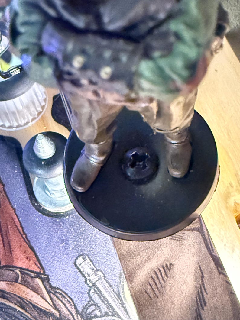 Puede incluir: Una figura en miniatura de un personaje con un atuendo de estilo militar, de pie sobre una base circular negra. La figura tiene una pintura detallada con tonos de verde, marr&oacute;n y plata. La base tiene un tornillo en el centro.