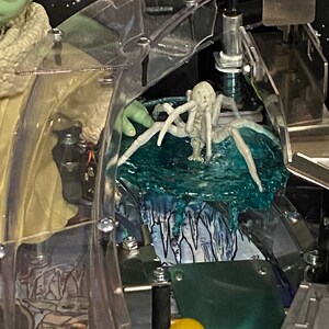 May include: A pinball machine playfield detail featuring a light green creature with large dark eyes and a textured beige robe, positioned next to a pale, multi-legged spider-like figure resting on a translucent blue, textured ice-like surface.