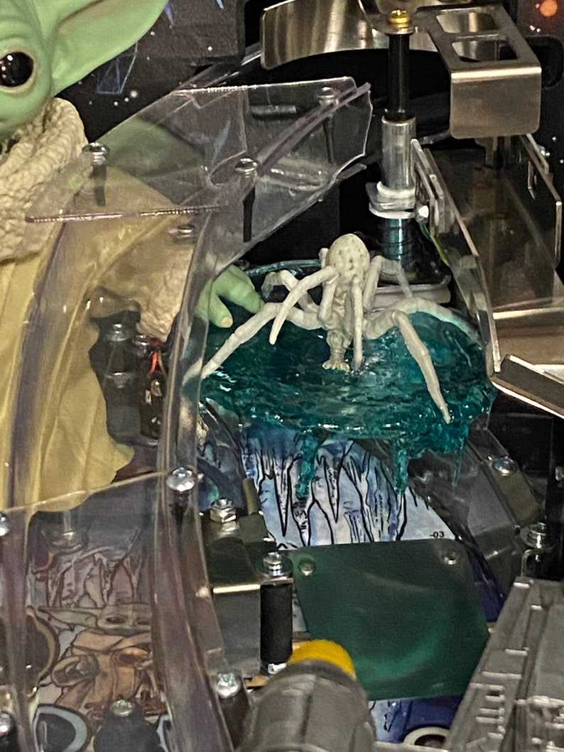May include: A pinball machine playfield detail featuring a light green creature with large dark eyes and a textured beige robe, positioned next to a pale, multi-legged spider-like figure resting on a translucent blue, textured ice-like surface.