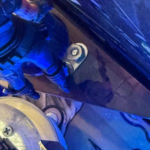 Puede incluir: Primer plano de una bisagra met&aacute;lica plateada fijada a una superficie azul oscuro. Una peque&ntilde;a figura detallada en azul y negro es parcialmente visible, a&ntilde;adiendo un toque de detalle a la escena. La imagen est&aacute; bien iluminada.