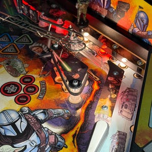 Puede incluir: Vista detallada de una m&aacute;quina de pinball con tem&aacute;tica de Star Wars. El campo de juego muestra ilustraciones vibrantes de personajes y escenas de la serie, con una peque&ntilde;a figura de un personaje. La m&aacute;quina tiene varias luces y componentes mec&aacute;nicos.