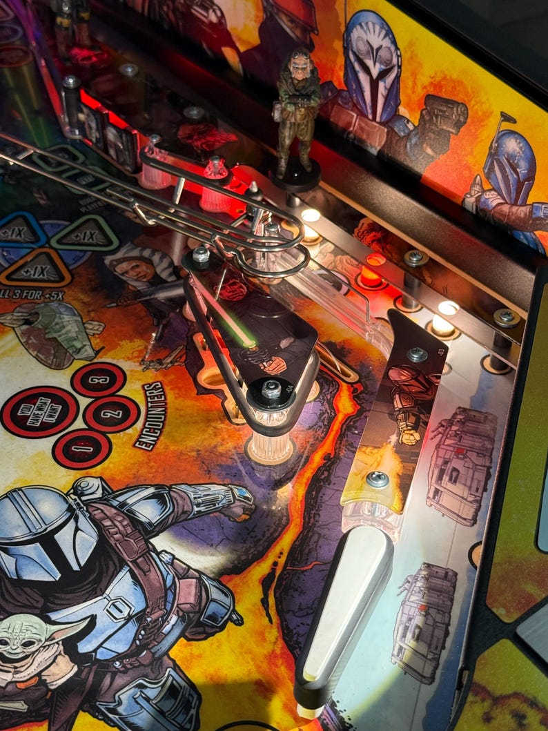 Puede incluir: Vista detallada de una m&aacute;quina de pinball con tem&aacute;tica de Star Wars. El campo de juego muestra ilustraciones vibrantes de personajes y escenas de la serie, con una peque&ntilde;a figura de un personaje. La m&aacute;quina tiene varias luces y componentes mec&aacute;nicos.