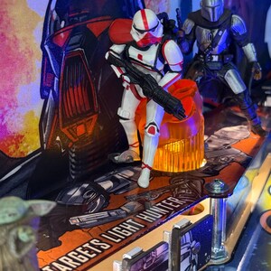 Puede incluir: Una m&aacute;quina de pinball con tem&aacute;tica de Star Wars con una figura de Stormtrooper blanca y roja sosteniendo un bl&aacute;ster negro, y una figura de Mandaloriano plateado. El campo de juego tiene el texto "TARGETS LIGHT HUNTER". El fondo presenta una gran ilustraci&oacute;n de un casco.