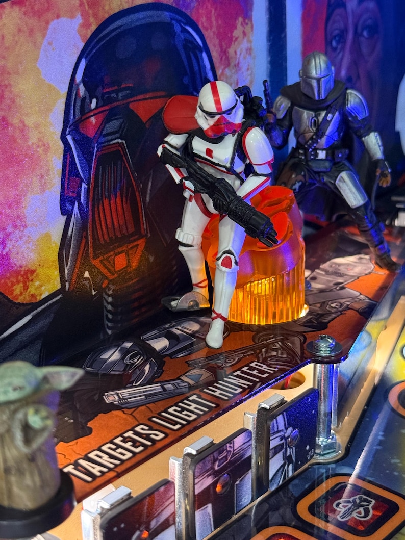 Puede incluir: Una m&aacute;quina de pinball con tem&aacute;tica de Star Wars con una figura de Stormtrooper blanca y roja sosteniendo un bl&aacute;ster negro, y una figura de Mandaloriano plateado. El campo de juego tiene el texto "TARGETS LIGHT HUNTER". El fondo presenta una gran ilustraci&oacute;n de un casco.