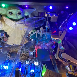 May include: A pinball machine playfield featuring a green creature figurine with large eyes and a cream-colored robe, a translucent blue spider-like figure, clear plastic ramps, metal hardware, and blue LED lights illuminating the game surface.