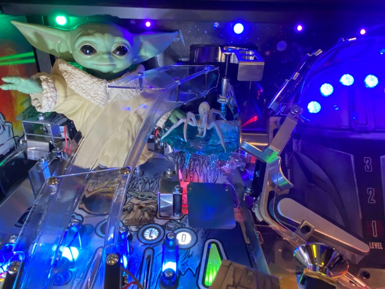 May include: A pinball machine playfield featuring a green creature figurine with large eyes and a cream-colored robe, a translucent blue spider-like figure, clear plastic ramps, metal hardware, and blue LED lights illuminating the game surface.