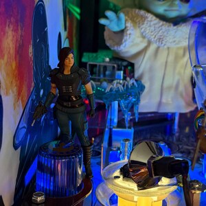 Puede incluir: Una m&aacute;quina de pinball con personajes de Star Wars. Una figura de Cara Dune se encuentra sobre un poste azul y plateado. El Ni&ntilde;o, tambi&eacute;n conocido como Baby Yoda, est&aacute; en el fondo. La m&aacute;quina tiene luces y obras de arte coloridas.
