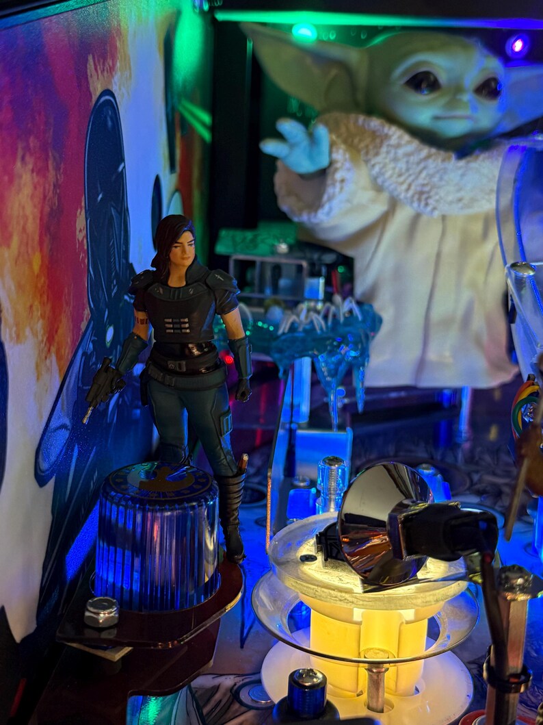 Puede incluir: Una m&aacute;quina de pinball con personajes de Star Wars. Una figura de Cara Dune se encuentra sobre un poste azul y plateado. El Ni&ntilde;o, tambi&eacute;n conocido como Baby Yoda, est&aacute; en el fondo. La m&aacute;quina tiene luces y obras de arte coloridas.